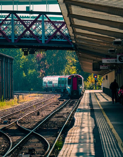 A modern commuter train with a sleek silver and red exterior is stationary at Enfield Lock station platform. The train has a smooth, metallic surface reflecting ambient light, with closed windows and 